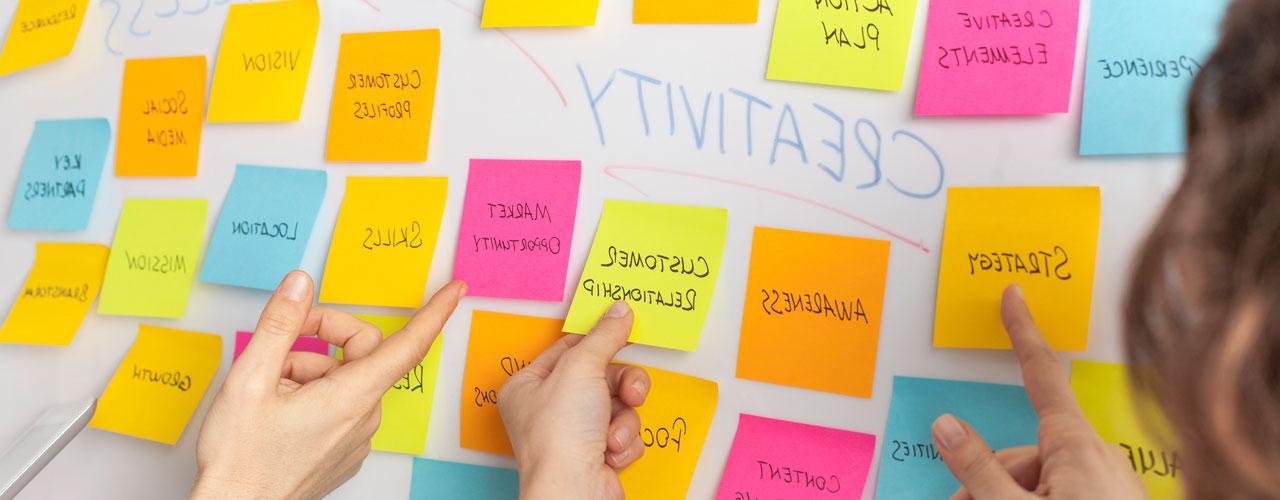 Hands pointing at a series of creativity-related sticky notes on a whiteboard.