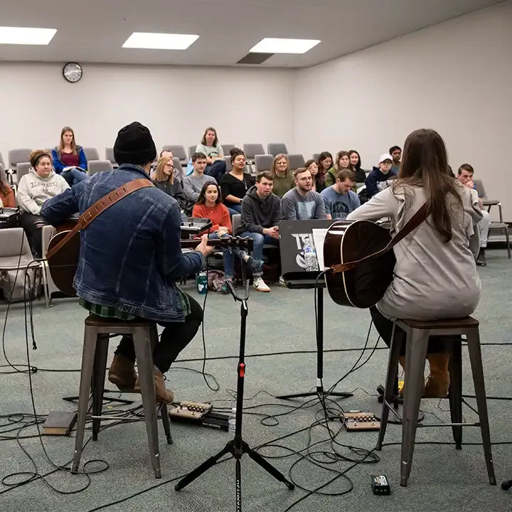 Two people playing guitars in room of people